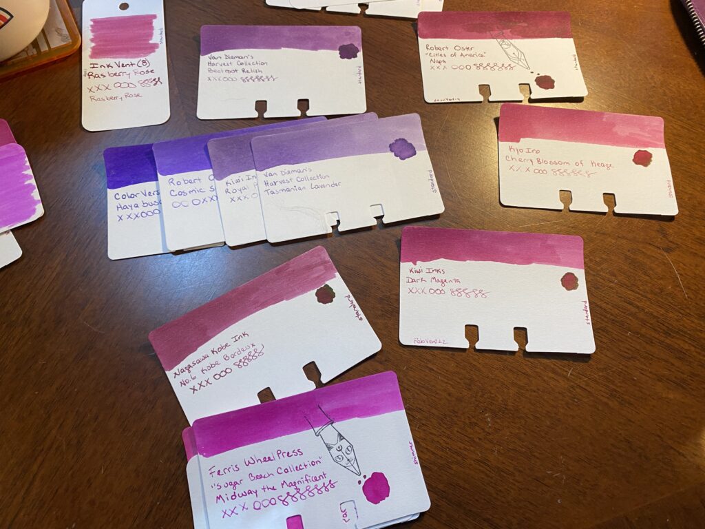 Lots of sample cards on a wooden desk. The purples have been narrowed down to 4 options, and there are magenta sample cards placed around it in smaller groupings or singly. 
