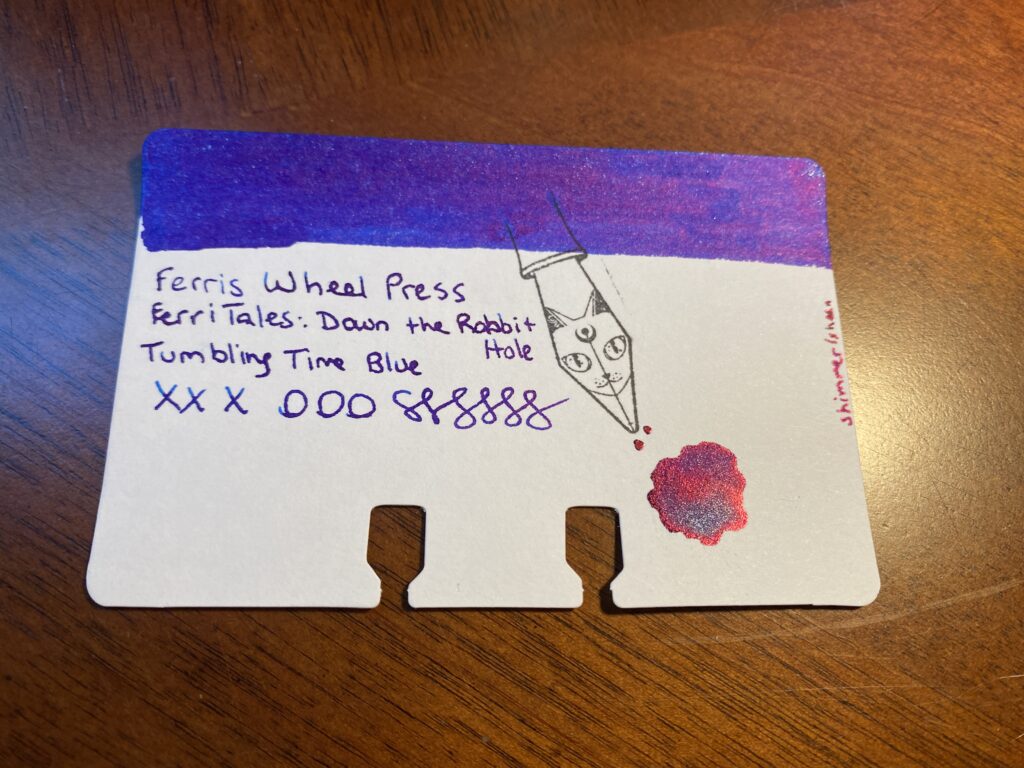 A single sample card on a wooden table of Ferris Wheel Press 
FerriTales: Down the Rabbit Hole
Tumbling Time Blue. There is a stamp on the card that looks like a fountain pen nib with a cats face engraved on it. 