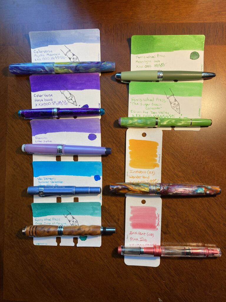 There are nine pens and ink sample cards laid out on a wooden table in two rows. The first row starts with a dark purple, then a lighter purple, then a bright blue, then a teal. The second row starts with a dark green, then a lighter green, then an orange and lastly a pink.The names of the pens and inks are listed below the photo.