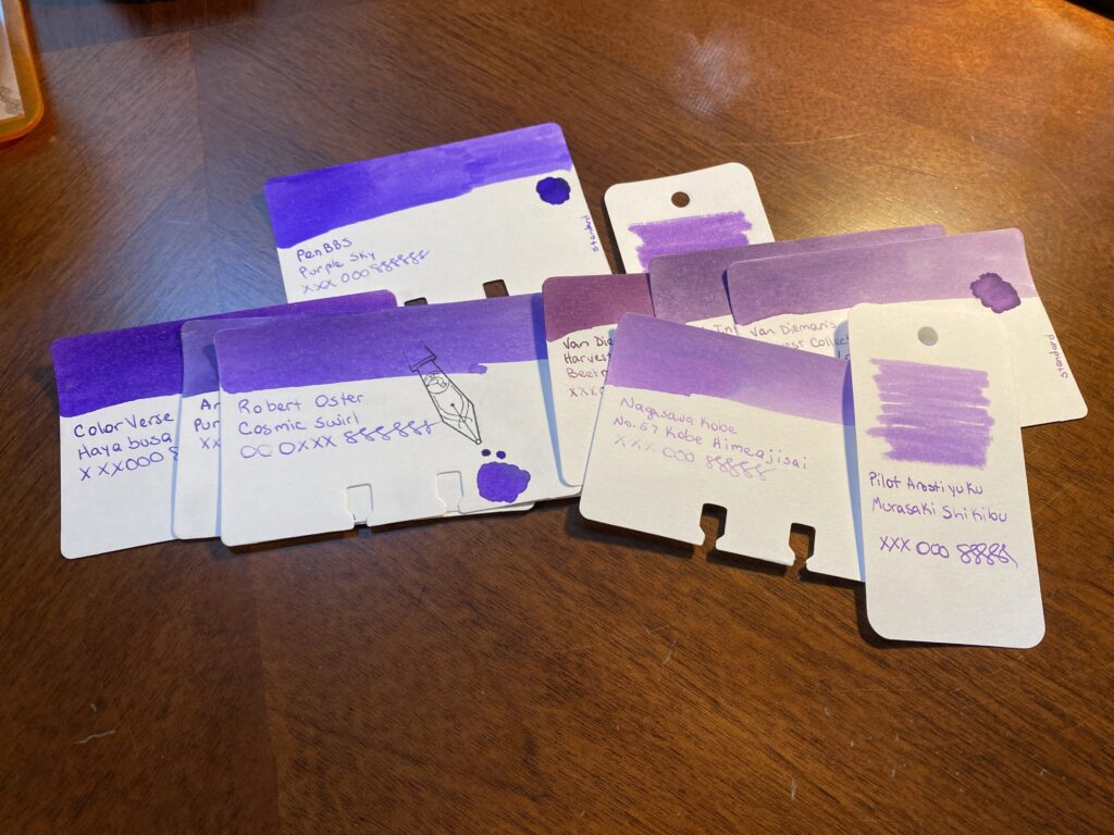 Pile of sample cards of purple inks, ranging from cool purples to warm purples. They are overlapping so the writing on them is close to each other. 