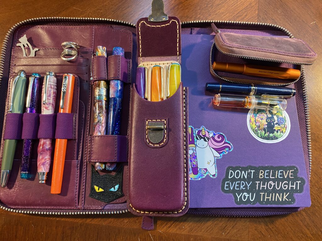 A purple folio with space on the right for a notebook. The one there is a more blue purple, with three stickers on it. One in the bottom right says “Don’t believe every thought you think.” And to the right is a unicorn with pink hair and sunglasses holding a sparkly fountain pen the same height as the unicorn. And above those is a lightbulb with a black cat and a garden inside.
On the right side of the portfolio is 4 card slots, and 6 pens. The card slots are holding a grey cat paper clip, and two ring splints. 
Set on top of the portfolio in the center is a purple external pen case that has space for 3 pens. And on the top right corner of the notebook is a smaller purple case, one pen rests inside, and 2 pens are set below it. 