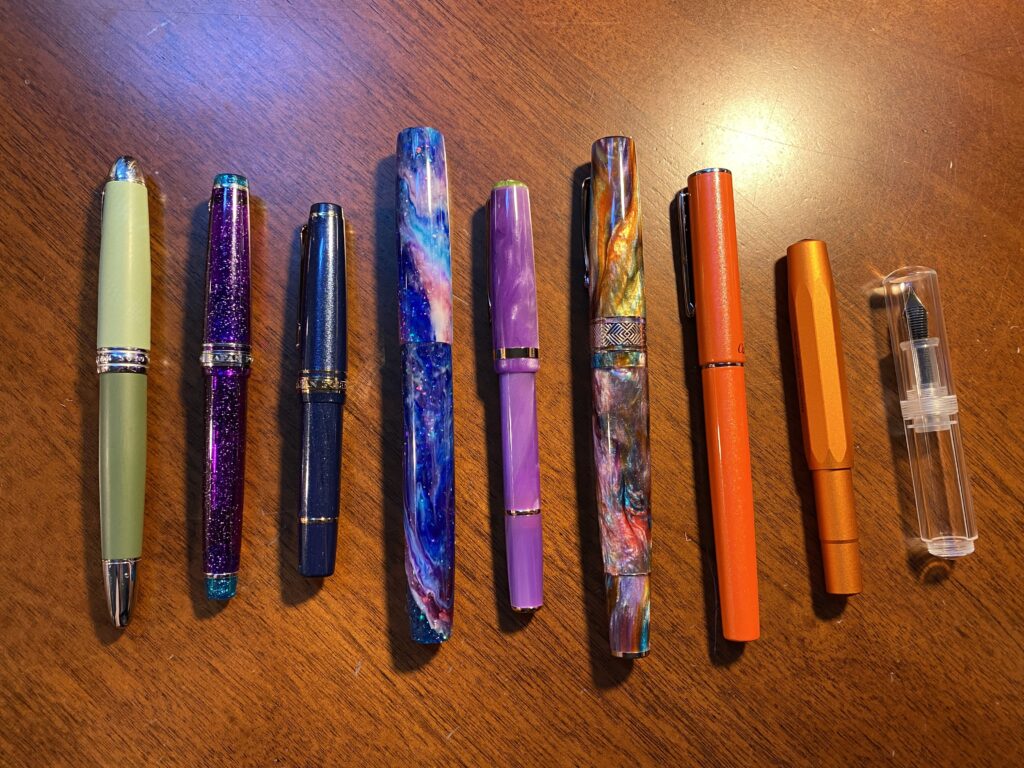 9 pens, starting from the left: two tone green, sparkly purple, sparkly blue, sparkly and swirly blue/purple/pinks, swirly purple, swirly and sparkly orange/purple/blue/green/yellow/pink, sparkly orange, a metallic orange, completely clear and tiny. 