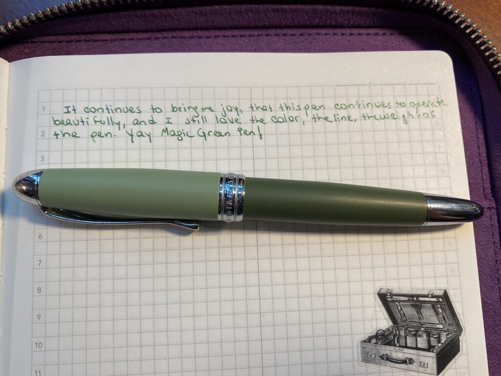 Notebook page with the following text: “It continues to bring me joy, that this pen continues to operate beautifully, and I still love the color, the line, the wright of the pen. Yay Magic Green Pen!” In a dark green ink with silver shimmer. Below the text is a two tone green pen with silver hardware. Below that is a sticker of an old Victorian suitcase. 