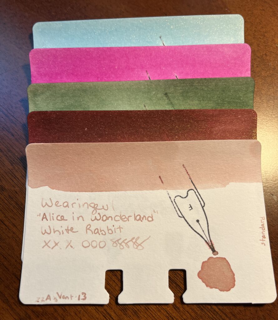 5 cards stacked on top of one another. The first four are only visible by the ink swabs along the time of the cards. The last card is fully visible and the text says:
Wearingeul 
“Alice in Wonderland”
White Rabbit
Xxx ooo sssss 
22AugVent-13 is written in the bottom left hand corner, and standard is written on the right edge of the card. 
The colors are a sparkly blue, bright pink, forest green, sparkly brownish maroon, and a pale tannish pink.