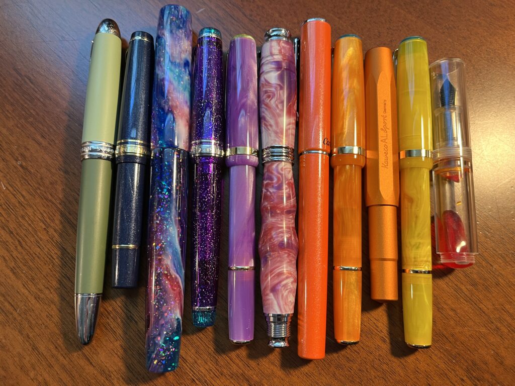 10 pens lined up horizontally, green, sparkle blue, blue/pink, sparkly purple, swirly purple, swirly magenta/pink/white, sparkly orange, swirly orange, metallic orange, swirly yellow, and clear with a sparkly orange ink visible.