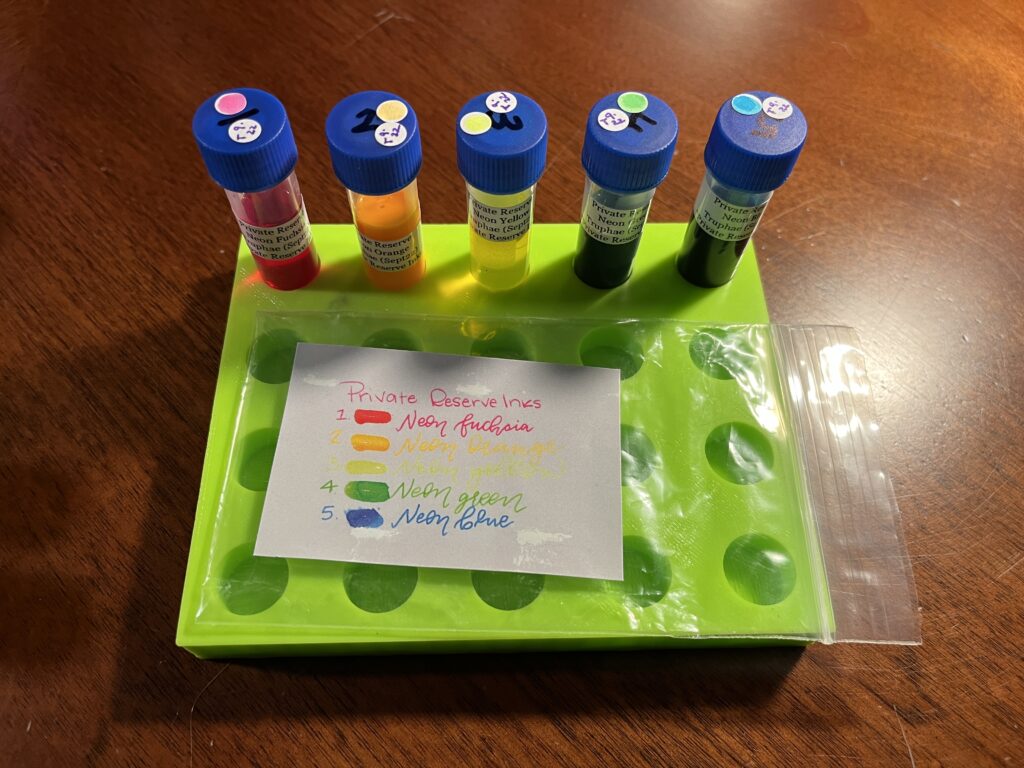 A lime green vial tray with 5 ink vials. Each one contains a brightly colored ink. An empty plastic bag with a label is resting in front of the vials. Each line is written in that color ink. It reads “Private Reserve Inks, 1 Neon Fuschia, 2 Neon Orange, 3 Neon Yello, 4 Neon Green, and 5 Neon Blue. 