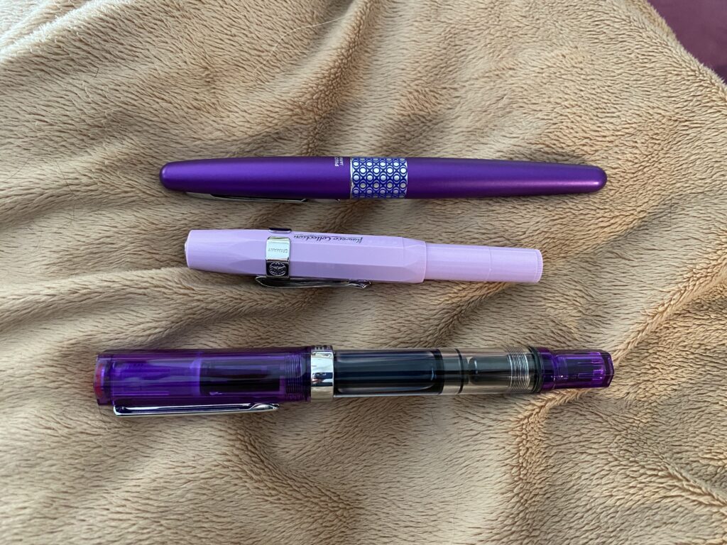 Three purple pens, the top is a purple Pilot Metropolitan, the second one is a light purple Kaweco Sport, and the last is a purple Twsbi Eco, all laying on a tan blanket.