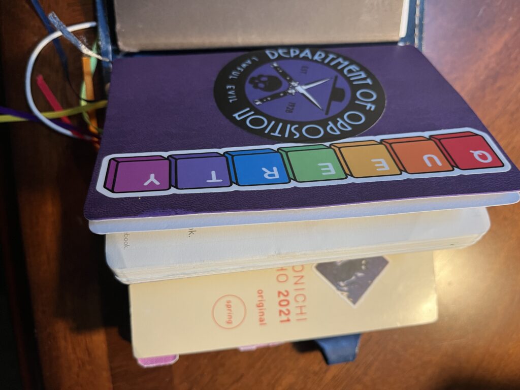 A notebook cover with three notebooks. Laying to the left is a brownish cover, in the middle is a purple cover with two sticks reading “Department of Opposition Lawful Evil” on one and “Queerty” on the other. Under the purple book is a Hobonichi Techo cover. 