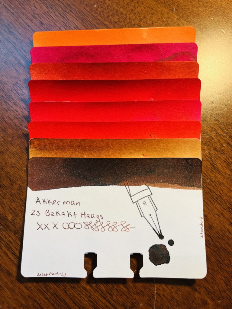 8 sample cards of an orange, different reds, and a couple of browns stacked on top of each other, only the color band at the top is visible on 7 of the cards. The last one is fully visible. It reads “Akkerman 23 Bekakt Haags, xxxooosssssss, 22SeptVent-23, standard” There is a stamp of a nib with a B on it, and a pool of the color used on the card. 