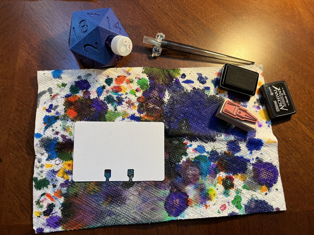 On a wooden desk are:
- paper towel which caught a ton of ink drops and spills 
- a blank white sample card
- a purple vial holder in the shape of a giant d20, which is holding a vial with a cap that has three white stickers on top. One is blank, one says “4” and the other says “9.22”
- a grey dip pen with a protector over the metal nib, and is resting on a glass pen holder
- a stamp ink pad with the lid taken off
- a stamp with the shape of a B nib