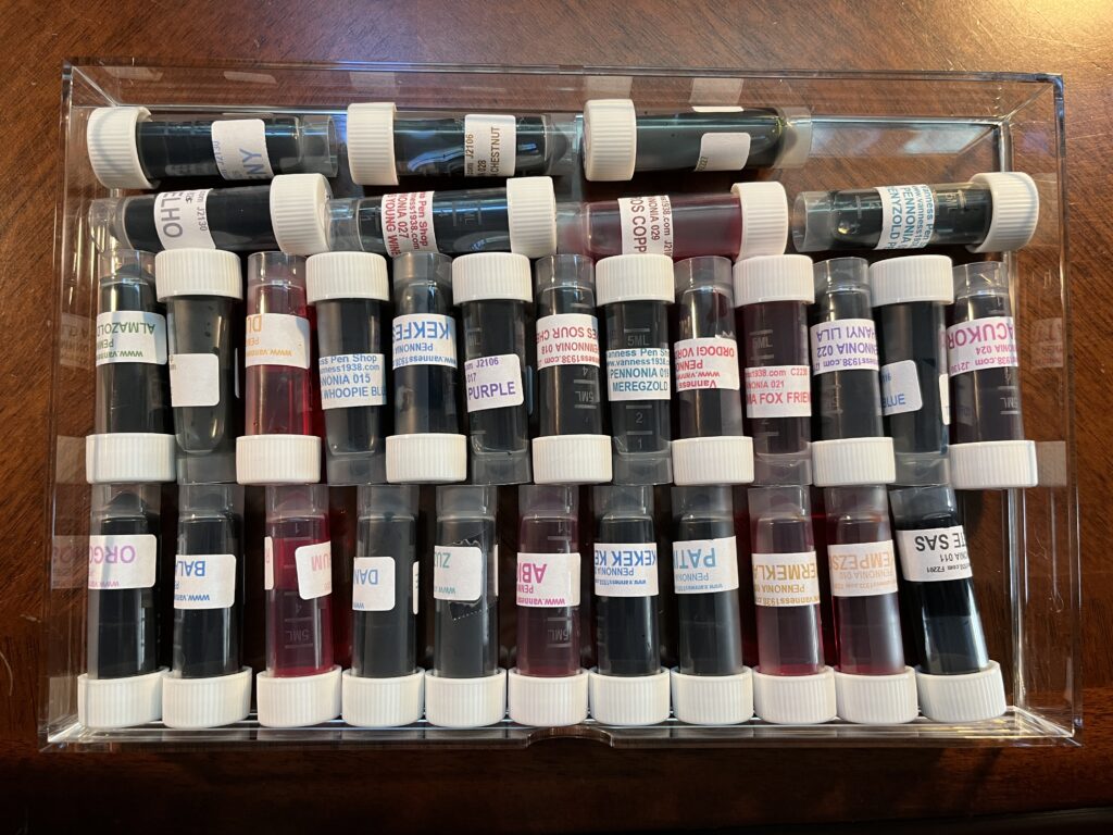 From the top down, a clear drawer with 31 ink vials, laying on their sides. Each vial has a white cap, and the inks are various colors. The labels are not all readable because they wrap around the vial, they are also all different colors, unsure if they match the ink witching the vial. 