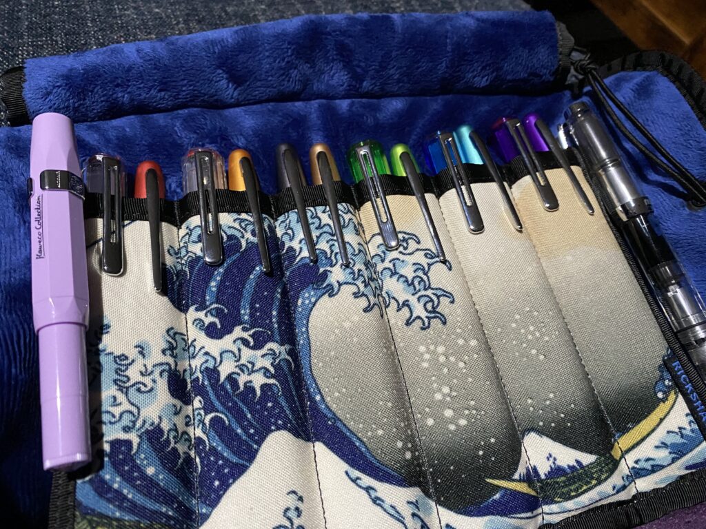 A pen holder with 14 pens. The pens are all the colors of the rainbows, and the pen case is a roll up with a classic wave pattern and the inside is a soft blue fabric. 