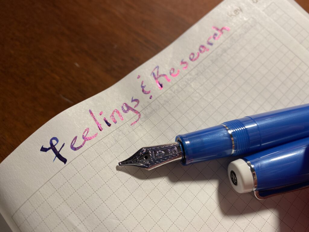 Notebook page with the text “Feelings & Research” in a blue ink with a purple sheen. There is an uncapped pen resting on the page, a blue resin with a white finial and a music nib.