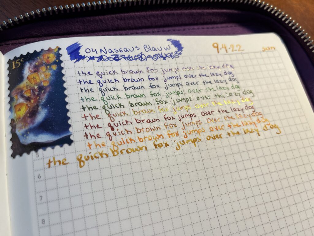 A notebook page - there is a sticker applied to the top left corner, it looks like a sample that has floating lantern art. To the right of the sticker is text reading “04 Nassaus Blauw” with scribbled marks to the left and underneath and the right side. In gold ink in the top right hand corner the text reads “9.4.22 Sun”
underneath is the text “the quick brown fox jumps over the lazy dog” written 10 times, in 10 different inks. The first is purple, the next two are different blues, the next two are different greens, a shiny red with gold sheen, a maroon, a orangey red with gold shimmer, a yellow orange, and a gold with gold shimmer. 