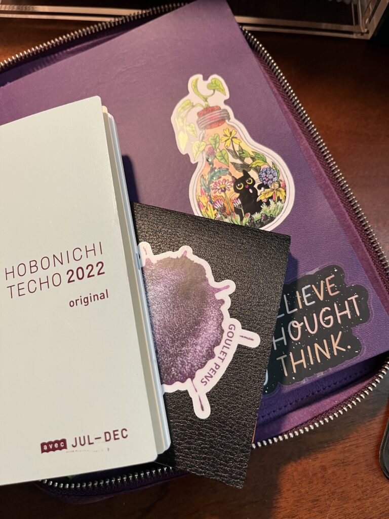 Three notebooks stacked on top of each other. The one on top is a Hobonichi Techo 2022 Original, underneath is a textured black notebook with a Goulet Pens purple ink splatter sticker on the cover, and the one on the bottom is purple, with 2 stickers visible. The top sticker is a lightbulbs with a garden and a black cat inside. And the bottom one is partially obscured, but the whole thing says, Don’t Believe Every Thought You Think.
