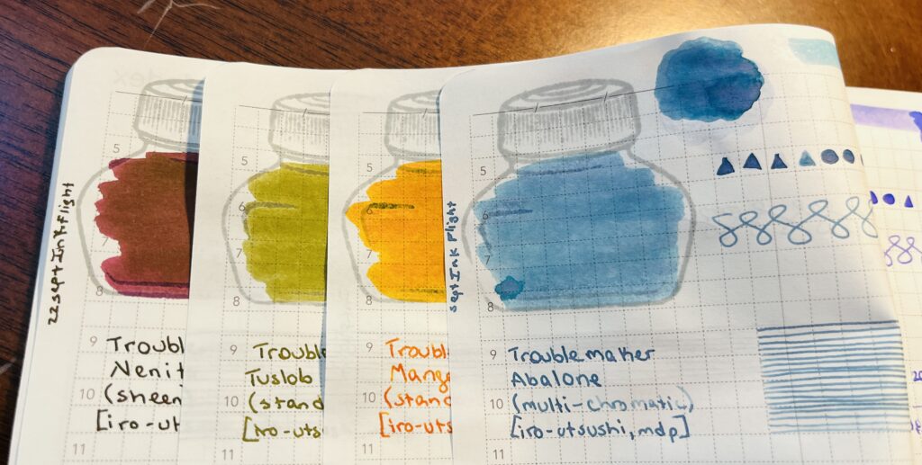 4 journal pages overlapping so each page can be seen enough to see the color. There is a maroon, a yellowish green, an orange, and a blue. The blue one is the only page mostly visible. There is an ink bottle stamp colored in with blue ink. The same blue ink was used to make triangle and circle shapes, some continuous s shapes, and straight lines. The text says “Troublemaker, Abalone, multichromatic, iro-utsushi, map”