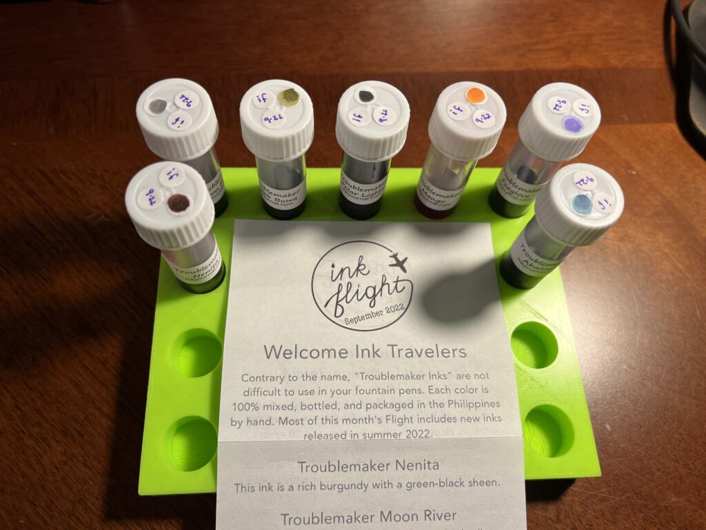 Lime green tray with seven vials of ink arranged around the paper with Ink Flight’s logo, September 2022, and text related to the Troublemaker’s inks. 