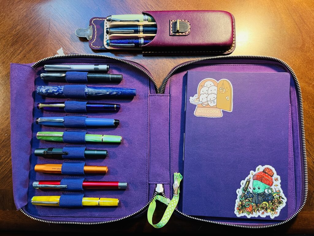 A purple notebook and pen holder. There are 10 pens on the left, 9 of them in loops, 1 loose. From top to bottom, a light silver, a darker grey, a blue, purple, white swirl, a dark purple, a light blue/purple, a light green, a dark green, an orange, a red, and a yellow. Above the notebook/pen case is a 3 pen pen case, with the Forever Purple and Blue pens, and the Magic Green pen. On the right of the notebook/pen holder is a purple notebook with two stickers on it. In the top left corner the sticker is 4 ghosts looking out of a front door and a welcome mat. In the bottom right hand corner is a green octopus with a red knit hat and a blue flannel coat. He has a fountain pen in two tentacles, a pumpkin in another, and a coffee cup in another. He is sitting in a pile of autumn leaves. 