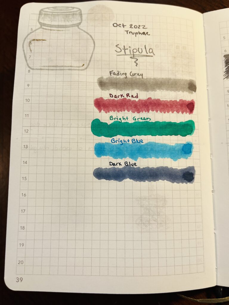 A single page of a journal. The text reads “Oct 2022 Truphae, Stipula” in a shiny grey. Each ink name is written with that ink color, and has a smudged line underneath of the same color. “Fading Grey, Dark Red, Bright Green, Bright Blue, Dark Blue.” 