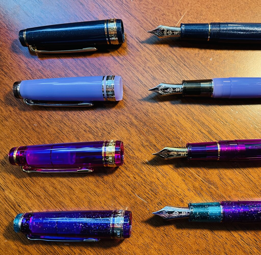 Four Sailor Pro Gear Slim pens, laid out horizontally with their caps off and nibs pointed to the left. The top one is sparkly blue, then a blue-purple, a darker purple and a sparkly purple. 