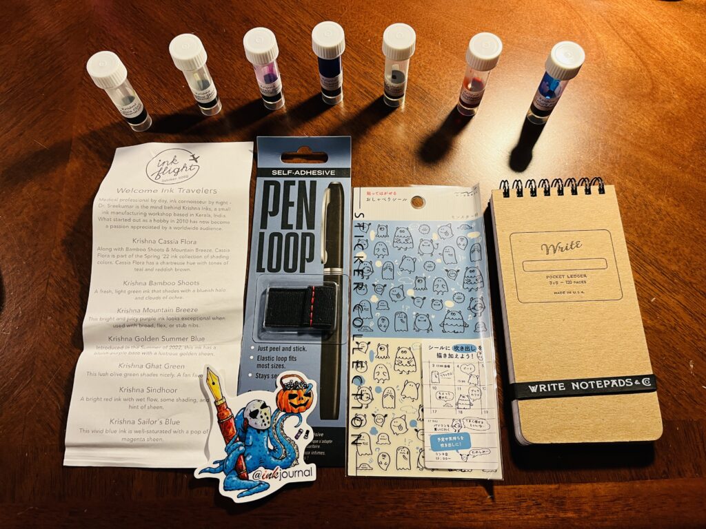 7 ink vials lined up horizontally, underneath from the left to write: the message Ink Flight sends about the box, a pen loop, an octopus sticker underneath, a sheet of ghost stickers, and a Write notepad.