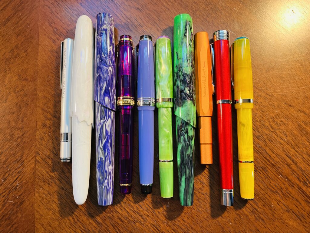 10 pens lined up side by side. There are a variety of types. There is a grey one, then white, then swirly purple, darker purple, lighter purple, light green, green and purple swirled, orange, red, and last a yellow. 