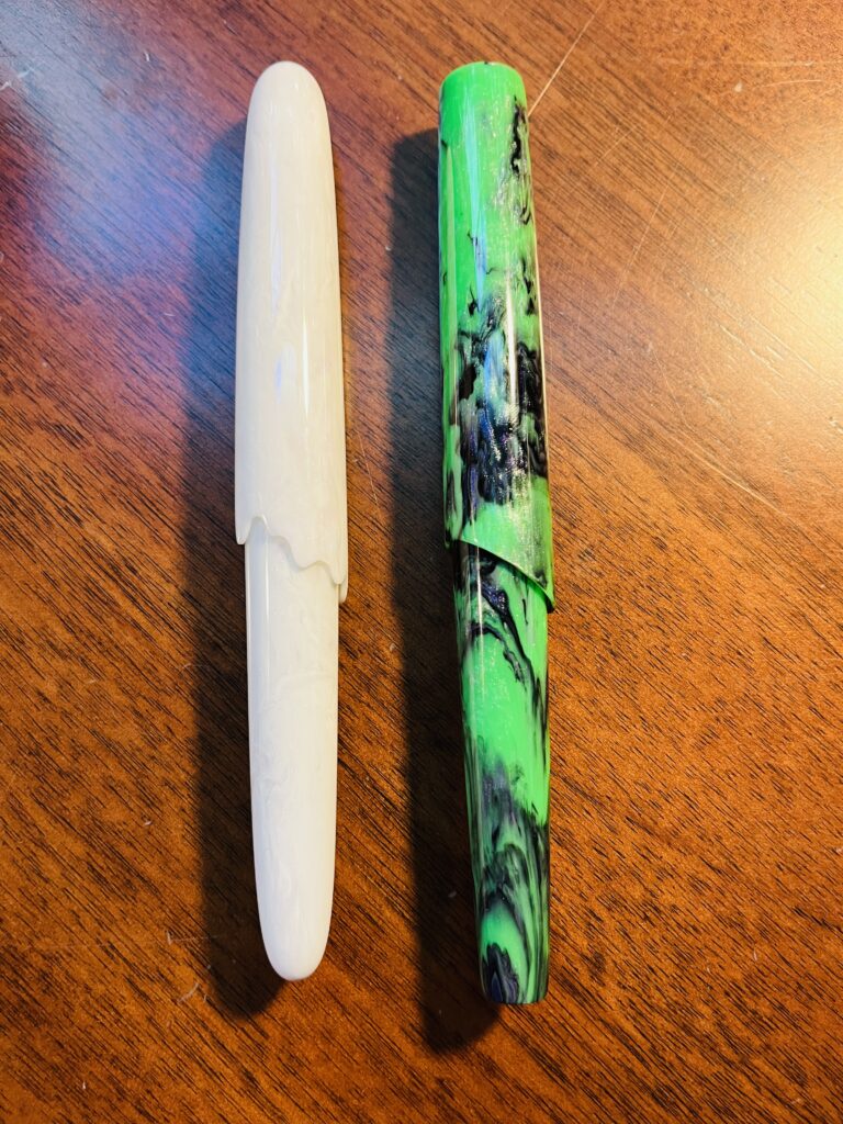 Two pens laid out vertically side by side. The one on the left is a swirly white, the edge of the cap has a swoopy edge instead of a straight edge, kind of looks like a ghost. The one on the right is a green and dark purple swirly, the top of the cap is a little more squared off than this model usually is, and the cap isn’t a straight line, but sort of angled. 