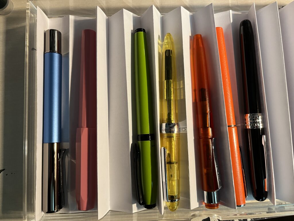 Top down view of 7 pens seated on paper dividers within a clear acrylic drawers. It looks kind of messy, the pens aren’t all equally visible, and some of the pleats have flattened.