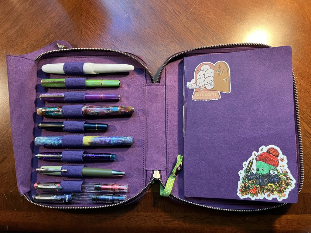 A purple notebook and pen case, with 10 pens on the left side, and a notebook on the right side. The notebook has two stickers, the one in the top left corner is 4 ghosts peeking out of a front door holding a cupcake, and with a welcome mat. The one in the bottom right hand corner is a green octopus with a red knit hat, and a blue flannel coat. He seems to be playing in a pile of autumn leaves and holding a fountain pen, a pumpkin, and a drink. 