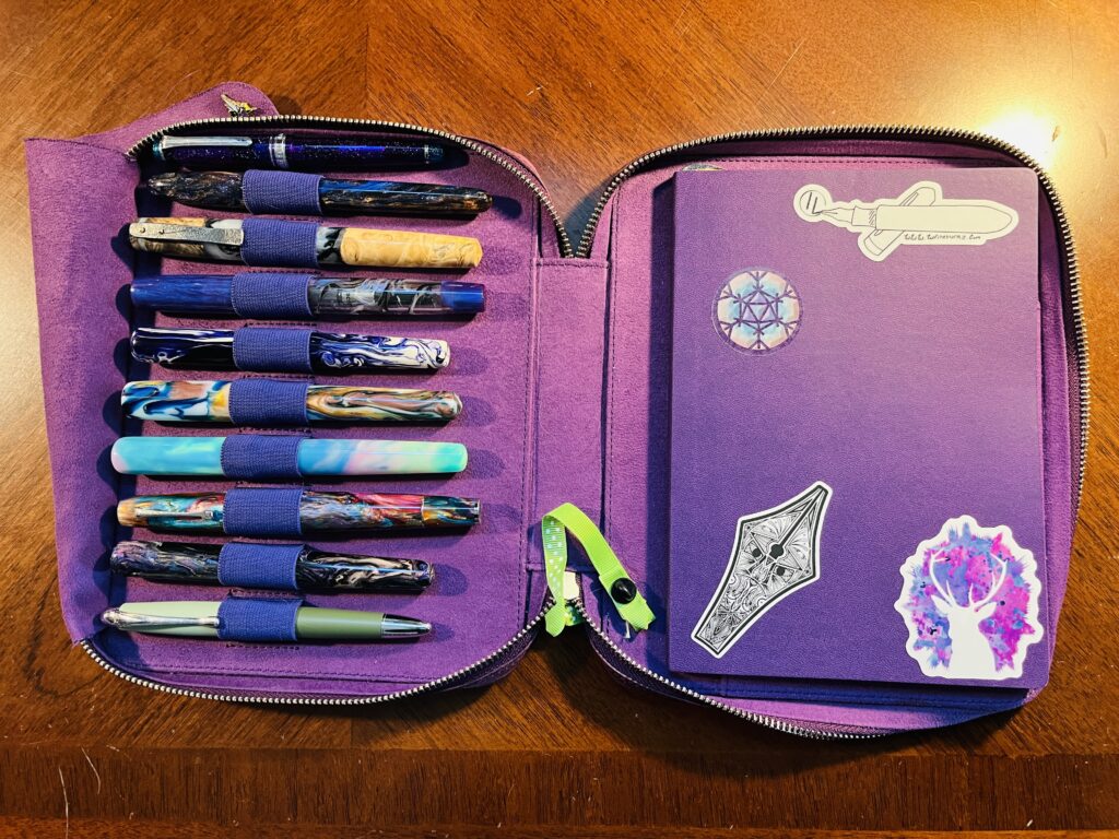 A pen and notebook portfolio. There are 10 pens, most made out of resin, some wood, and some metal types. The notebook is purple and has 4 stickers on it. One is a snowflake with a d20 in the center, then a nib with dancing etching, then a white fountain pen, and last a white silhouette of a deer with purple and pink background. 