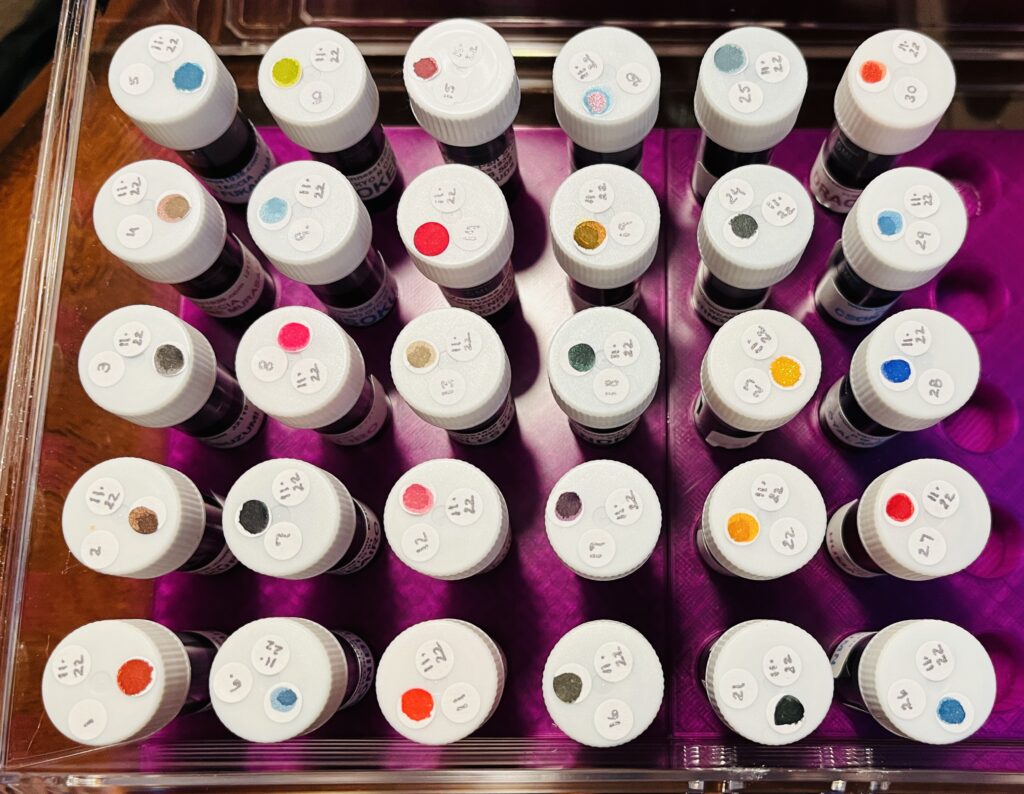30 ink vials from the top, only the white caps can be seen, each has 3 stickers. The vials are labeled 1-30, and each has the date 11.22, and each has a single color dot. 