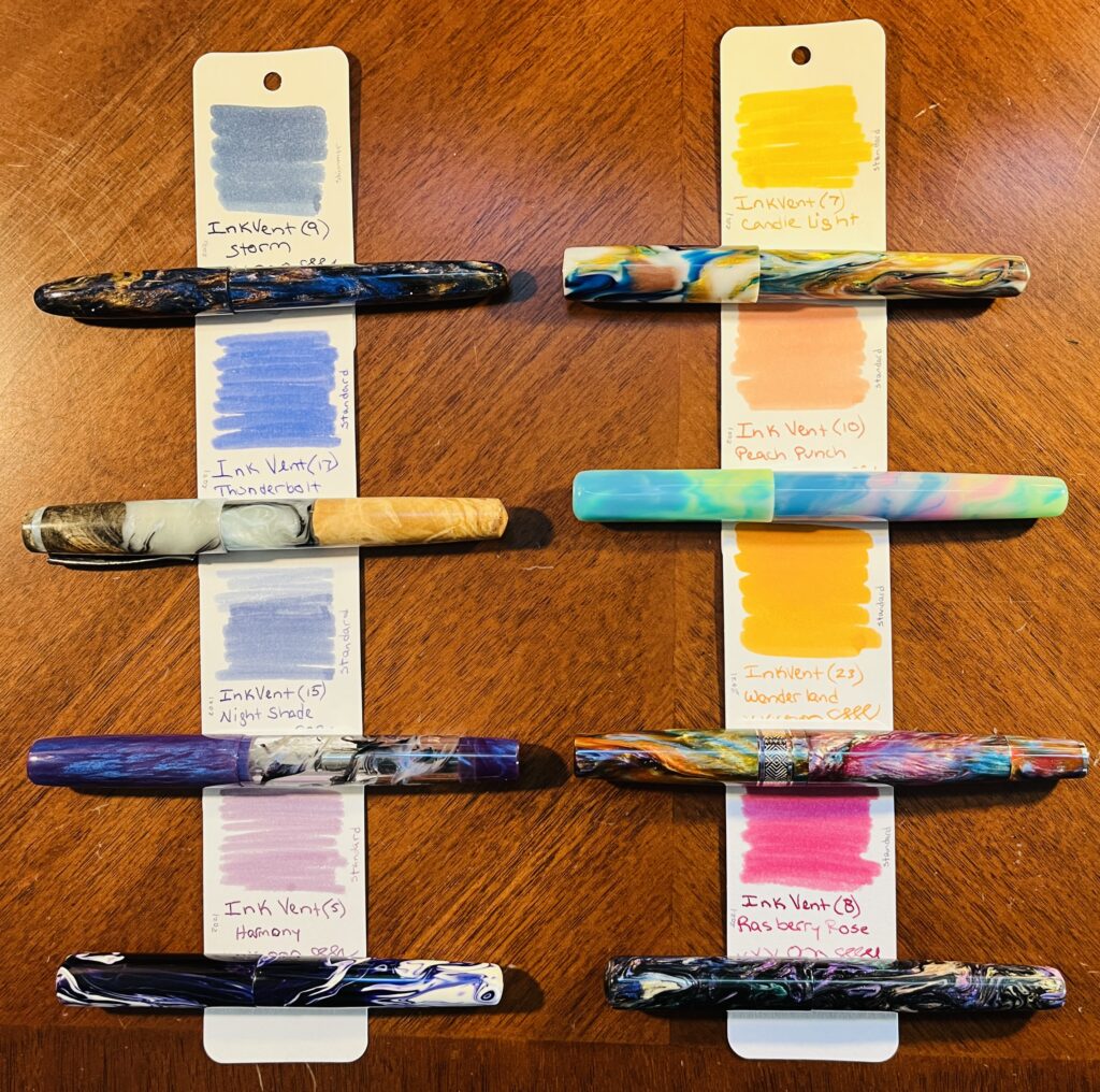 8 ink sample cards, each with a pen below where the name of the ink is written. The left column starts with a shimmer blue, then another brighter blue, then a blue purple, and a less saturated purple. The right column starts with a yellow, a sort of pinkish orange, a solid orange and what looks like a magenta but is more of a raspberry color. 