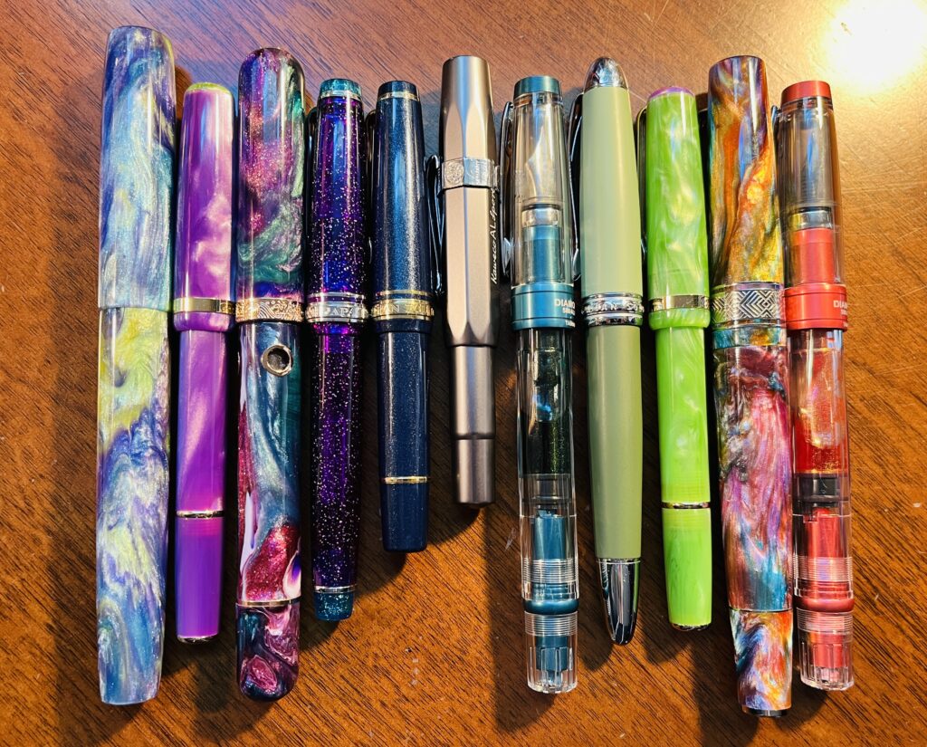 11 pens lined up horizontally, with the pens standing vertically. They are a combination of different colors and materials, shapes and lengths. 
