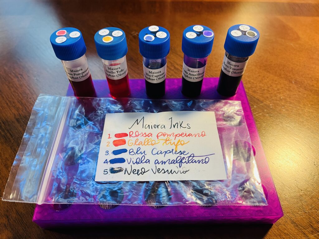 A purple tray with 5 ink vials lined up side by side and a plastic bag with the inks listed in handwriting that is difficult to read, each name written in the color of the ink. 