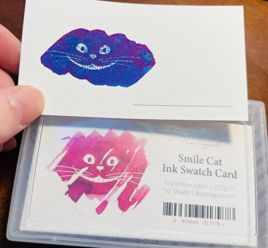 A container of Wearingeul Smile Cat Ink Swatch Cards, 200g’m, 5 sheets and a single swatch card above it with a blue ink with purple sheen and silver shimmer. 