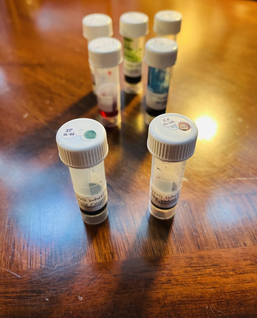7 sample vials with ink in them. They all have white lids, but only the front 2 have labeled dots on top. 